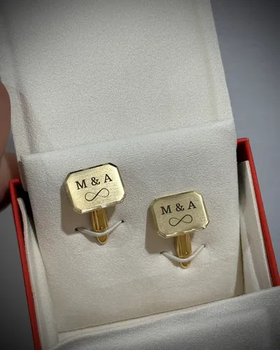 Zwei goldfarbene Manschettenknöpfe mit gravierten Initialen und Unendlichzeichen in einer Schatulle